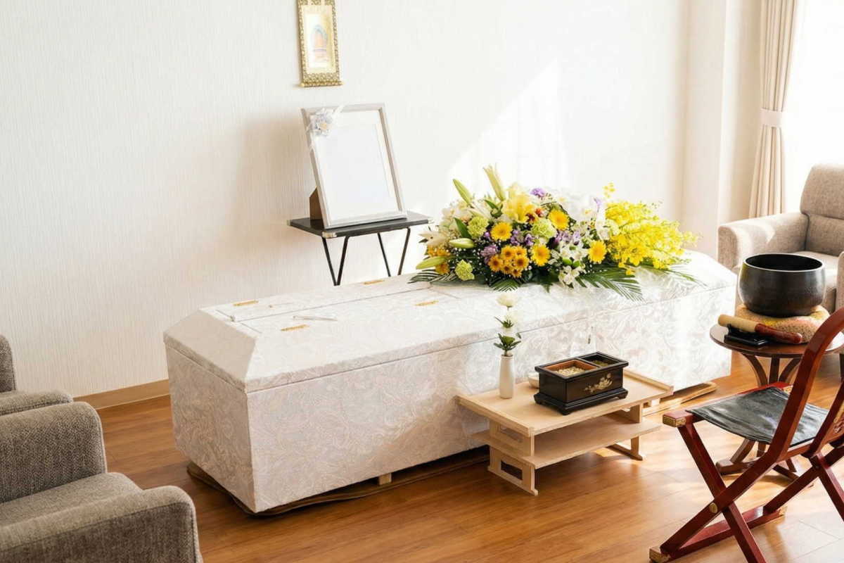 リビングのダイニングテーブルの奥に安置された白いお柩。お柩の上には色鮮やかな棺上花が飾られ、奥には小スペース用のシンプル祭壇が設けられた温かい自宅葬の風景